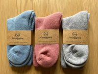 Three pairs of Moggans Cashmere Bed Socks, in blue, pink, and grey, each wrapped with a brown brand label, are neatly arranged side by side on a wooden surface. Made in Scotland.