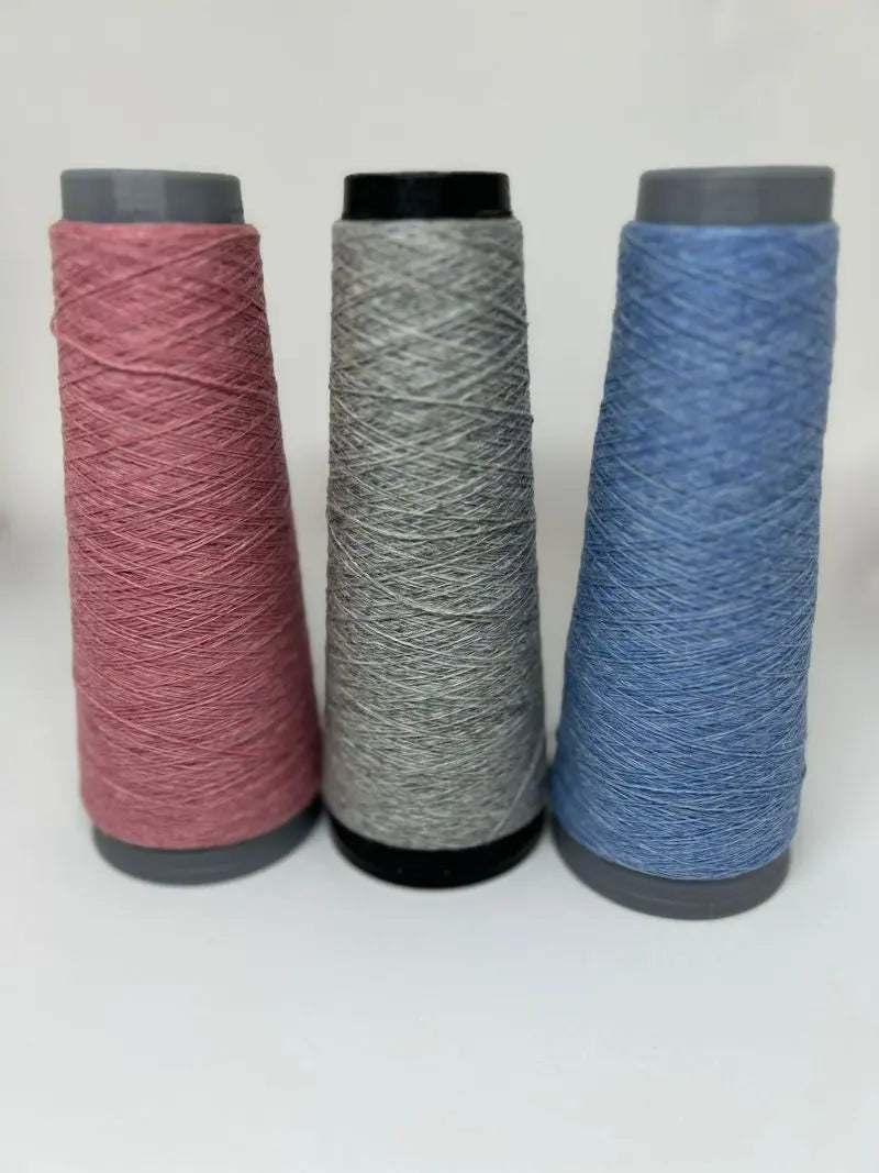 Three Moggans cashmere thread cones for bed socks—pink, gray, and blue—stand side by side on a white surface. Crafted in Scotland, they're perfect for knitting luxurious Cashmere Bed Socks.