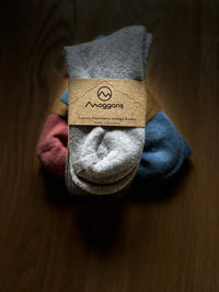 Three pairs of folded Moggans Cashmere Bed Socks—gray, blue, and pink—are wrapped in a brown paper label reading Cashmere Bed Socks - Made in Scotland by Moggans, arranged neatly on a dark wooden surface.