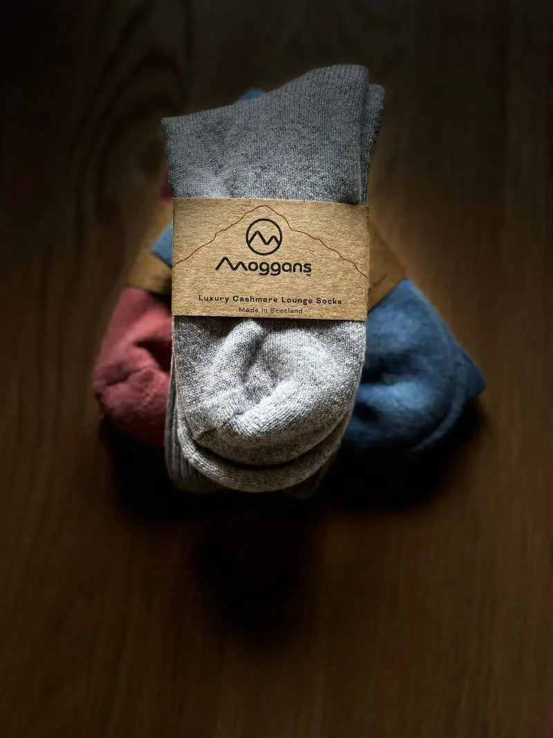 Three pairs of folded Moggans Cashmere Bed Socks—gray, blue, and pink—are wrapped in a brown paper label reading Cashmere Bed Socks - Made in Scotland by Moggans, arranged neatly on a dark wooden surface.