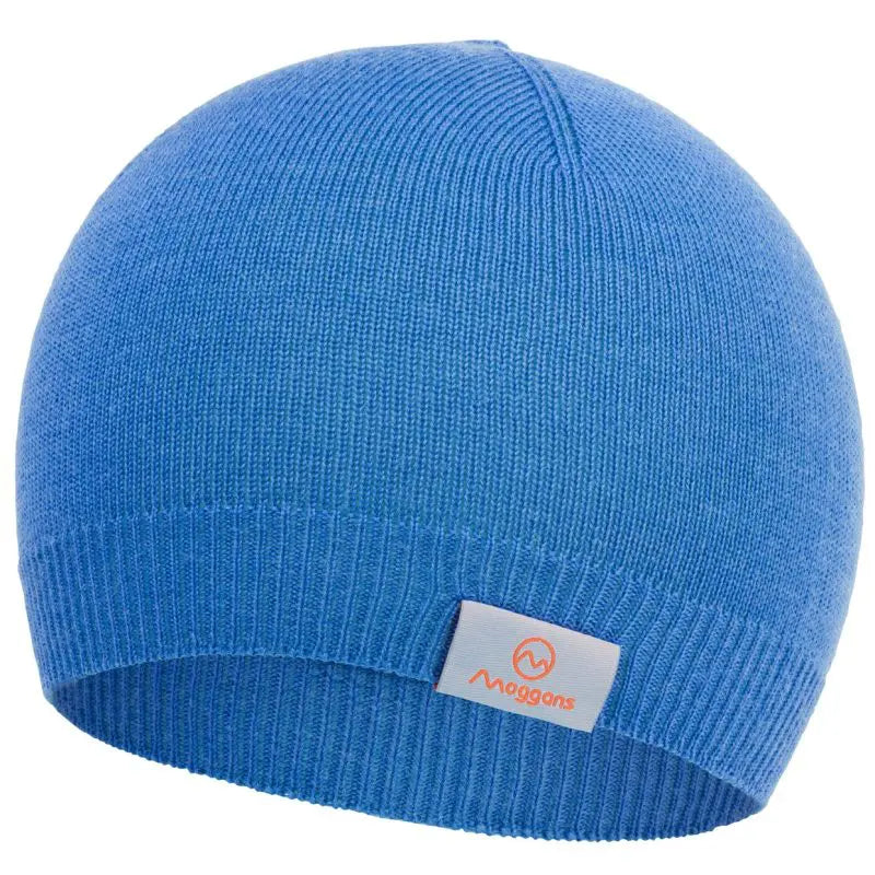 The 100% Merino Wool Beanie by Moggans is a plain blue knit hat made in Scotland from mulesing-free wool, featuring a ribbed edge and a small white tag with an orange logo and "Moggans" text on the front.
