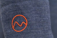 Close-up of the Moggans Merino Long Sleeve Baselayer with TENCEL™ in blue, featuring an orange, embroidered circular logo with a zigzag line resembling a mountain or wave.