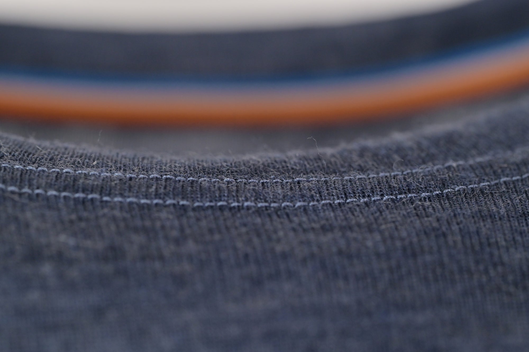 Close-up of the Moggans Merino Long Sleeve Baselayer with TENCEL™, showing dark blue textured Merino wool, visible stitching, and a hint of a shirt collar, with blurred orange and blue lines in the background.
