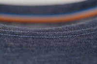 Close-up of the Moggans Merino Long Sleeve Baselayer with TENCEL™, showing dark blue textured Merino wool, visible stitching, and a hint of a shirt collar, with blurred orange and blue lines in the background.