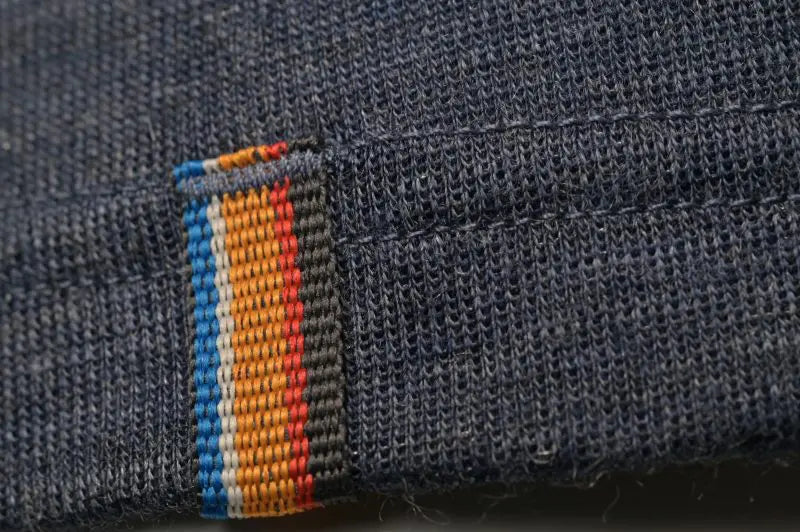 Close-up of the Moggans Merino Short Sleeve Baselayer with TENCEL™ shows its dark blue, moisture-wicking fabric and detailed stitching, plus a label stitched with four vertical stripes in blue, orange, yellow, and red.