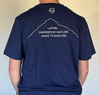 A person wears the Moggans Cotton Tee by Moggans, a navy blue Scotland t-shirt with a simple Buachaille Etive Mòr mountain outline and “INSPIRED BY NATURE MADE TO ENDURE” text below a circular logo on the back.