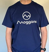 A person wearing a navy blue Moggans Cotton Tee featuring the brand’s white circular logo above Moggans on the front. The image focuses on the tee’s distinctive Scottish mountain-inspired style, with the persons head not visible.
